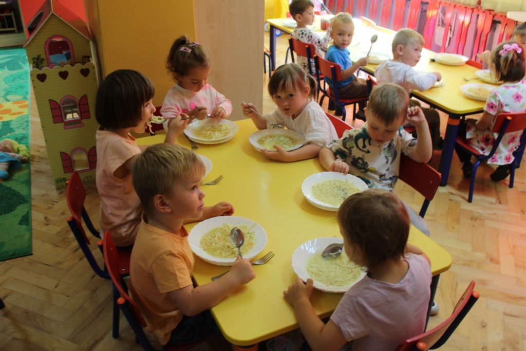 Szóstka dzieci siedzi przy stoliku i zjada rosół na drugim planie widzimy dzieci siedzące przy obiedzie
 