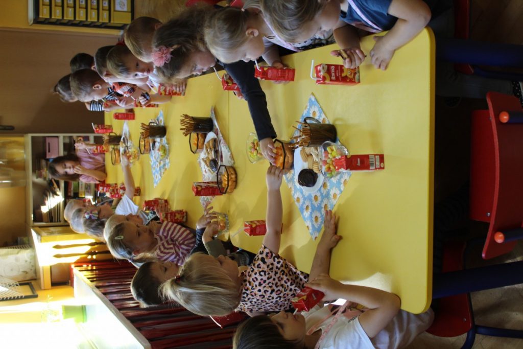 Grupa dzieci siedzi przy stole i częstuje się słodyczami i pije soczki z kartoników