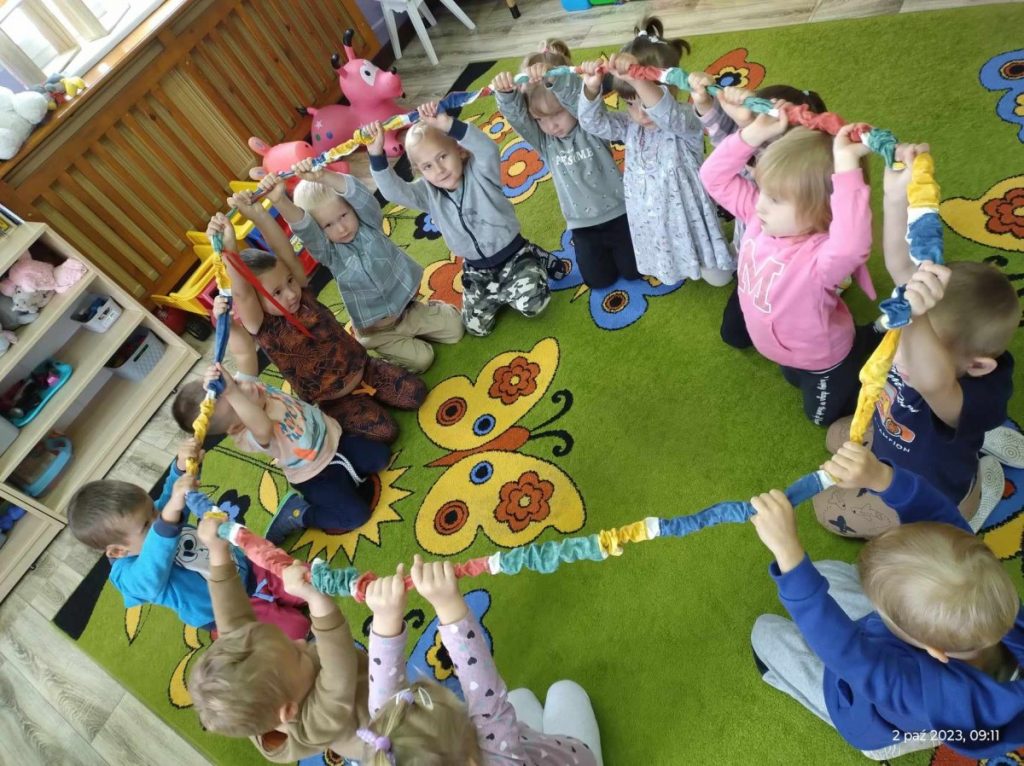 Na zdjęciu dzieci siedzą w kole na dywanie trzymając w rękach kolorową gumę.