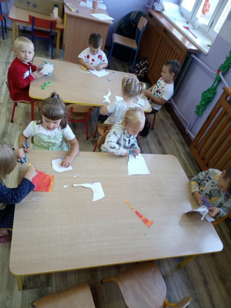 Na zdjęciu dzieci siedzą przy stolikach wycinają kształt kropki z kolorowego papieru.