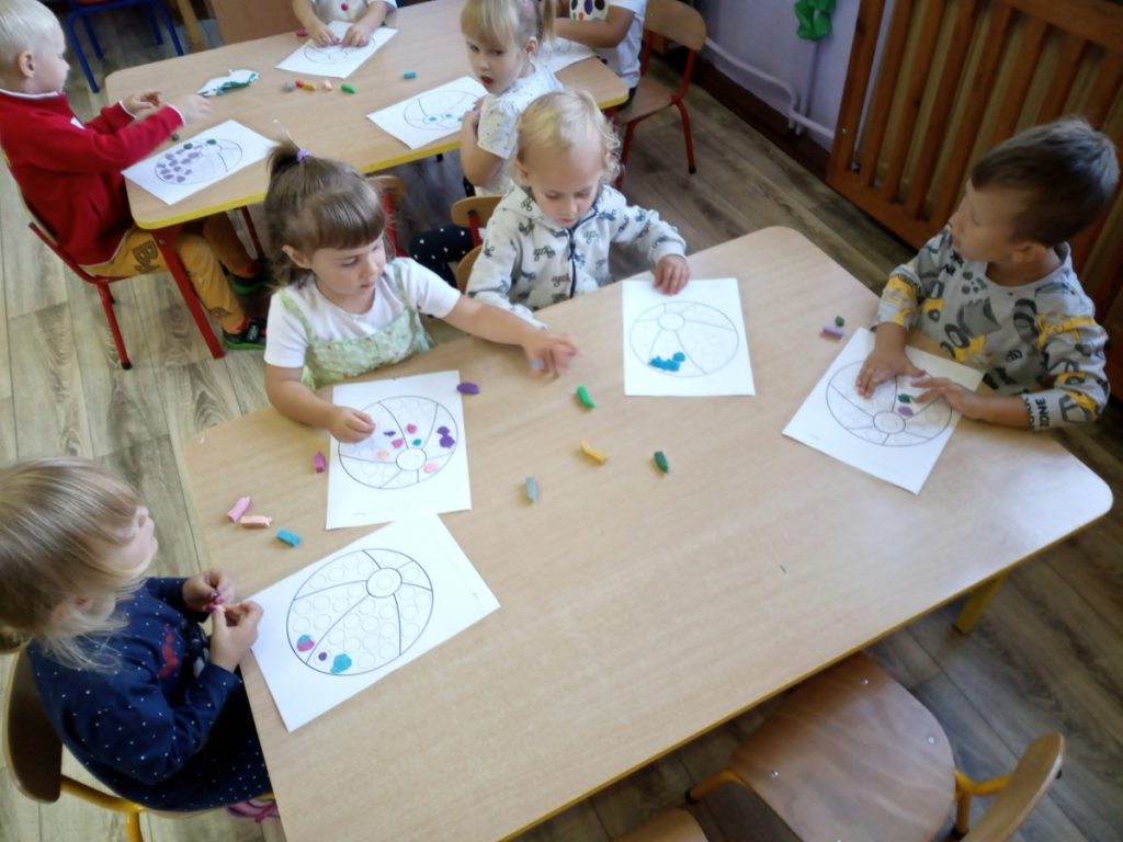 Na zdjęciu dzieci siedzą przy stolikach, wyklejają piłkę kolorowymi kulkami z plasteliny.