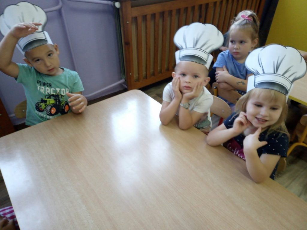 Na zdjęciu dzieci w czapkach kucharskich siedzą przy stoliku.