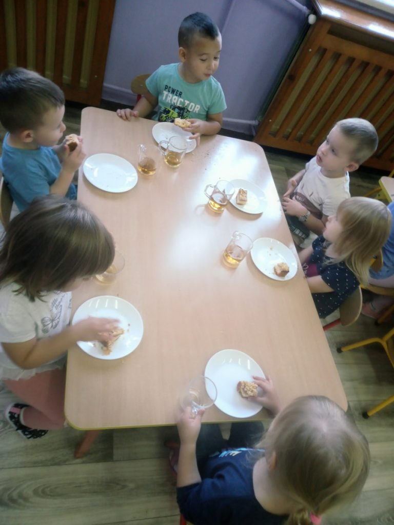 Na zdjęciu dzieci siedzą przy stoliku i jedzą upieczoną szarlotkę.