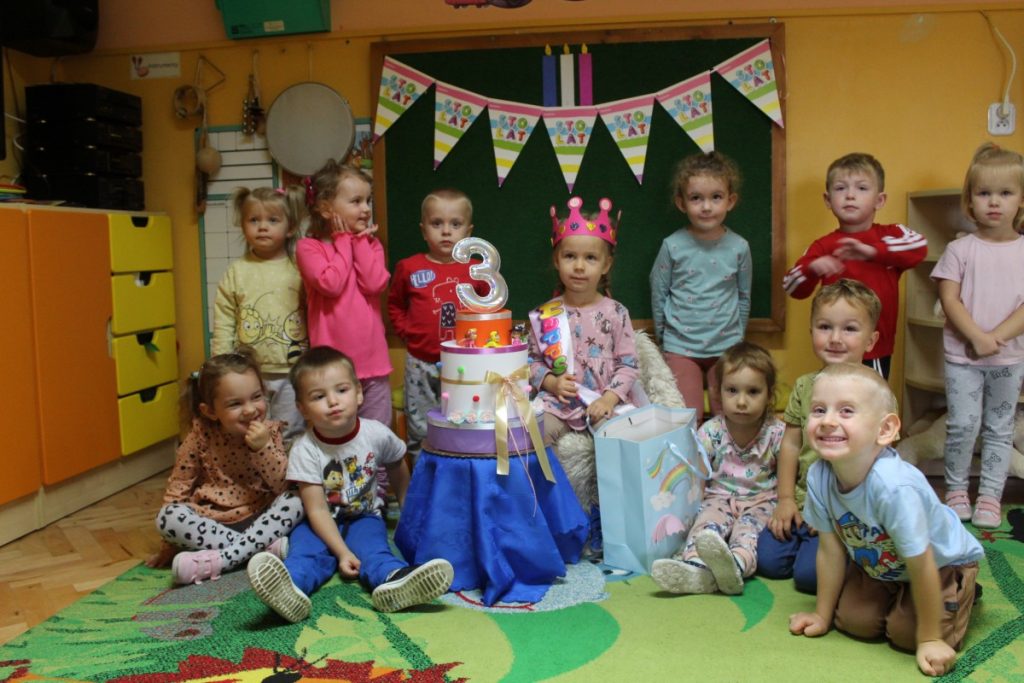 Dziewczynka w koronie obok tort z papieru z cyfrą 3 i dzieci siedzące i stojące