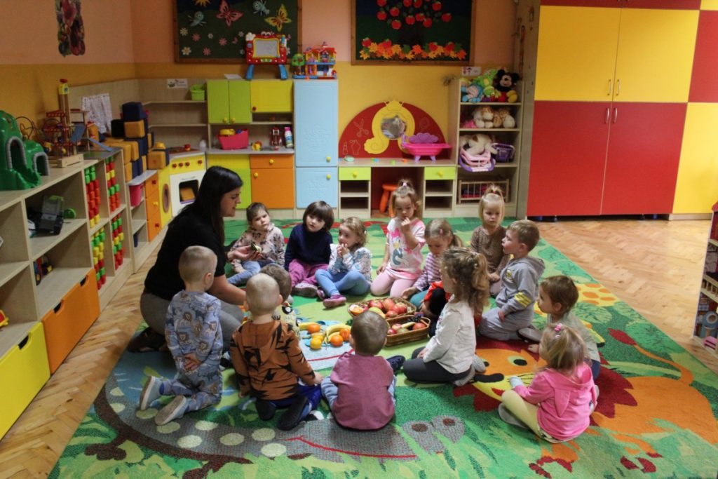 Dzieci na dywanie z nauczycielka siedzą w kręgu . Nauczycielka pokazuje owoce