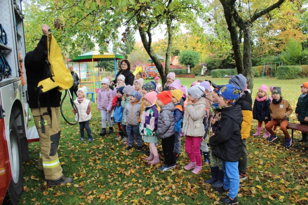 strażak pokazuje wyposażenie samochodu strażackiego a dzieci stoją na przeciwko