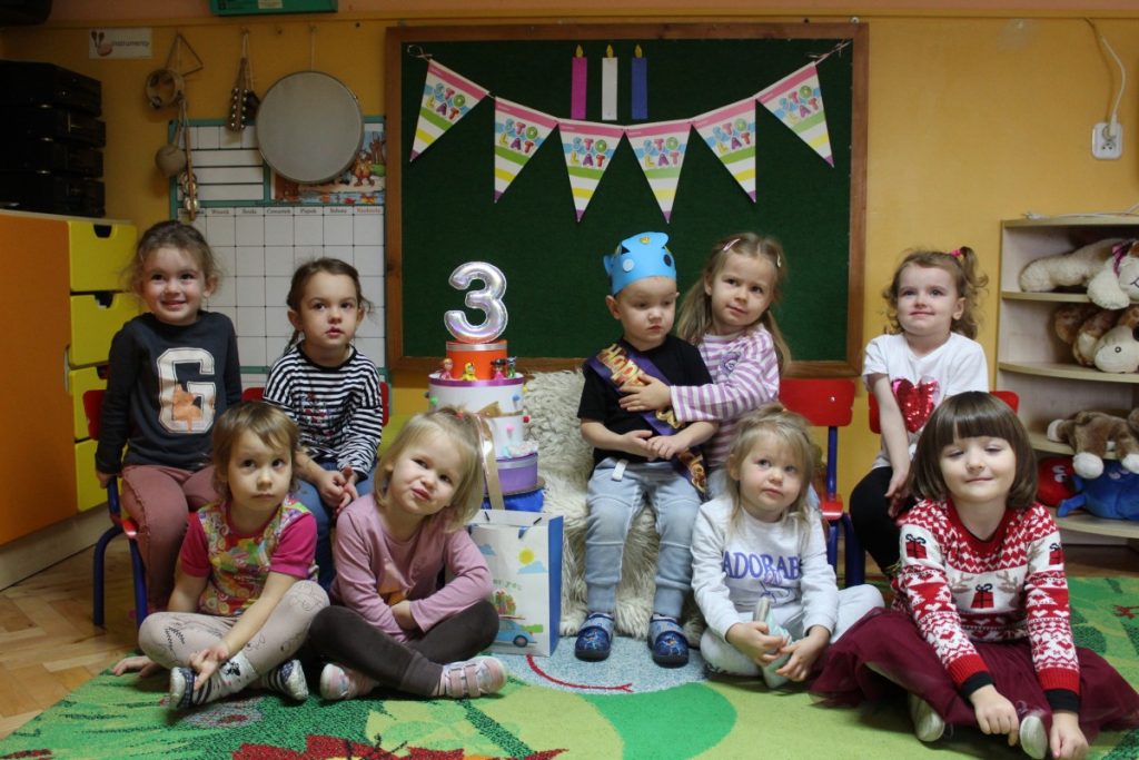Grupa dzieci w środku siedzi chłopiec w niebieskiej koronie a obok niego papierowy tort z cyfrą 3 