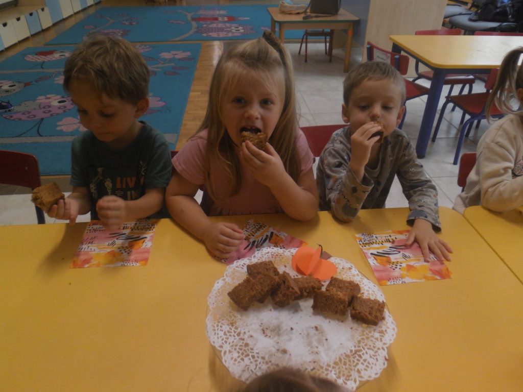 Na zdjęciu dzieci siedzą przy stolikach i zajadają ciasto z dyni