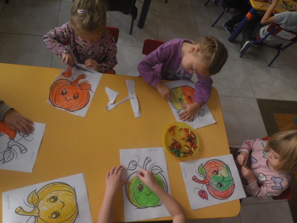 Na zdjęciu dzieci siedzą przy stolikach i malują kolorowankę dyni