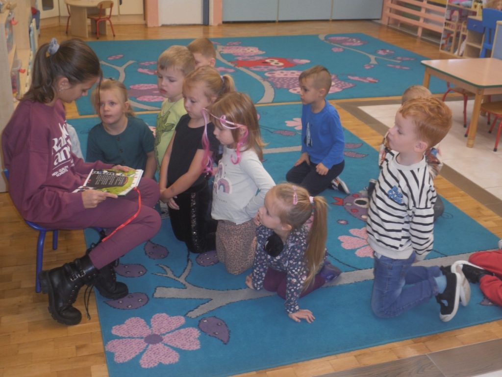 Na zdjęciu dzieci siedzą na dywanie i słuchają czytanej bajki