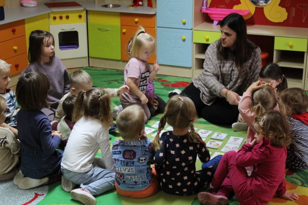 Dzieci wraz z nauczycielką siedzą
a na dywanie