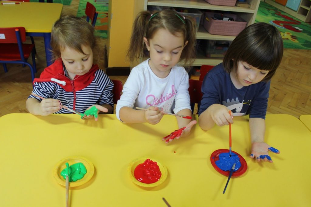trójka dzieci przy stoliku maluje pędzlem każde swoją dłoń