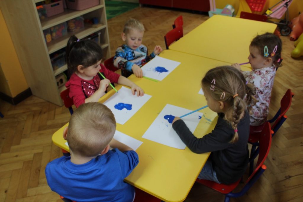 Dzieci przy stolikach dmuchają przez rurki na niebieskiego kleksa z farby