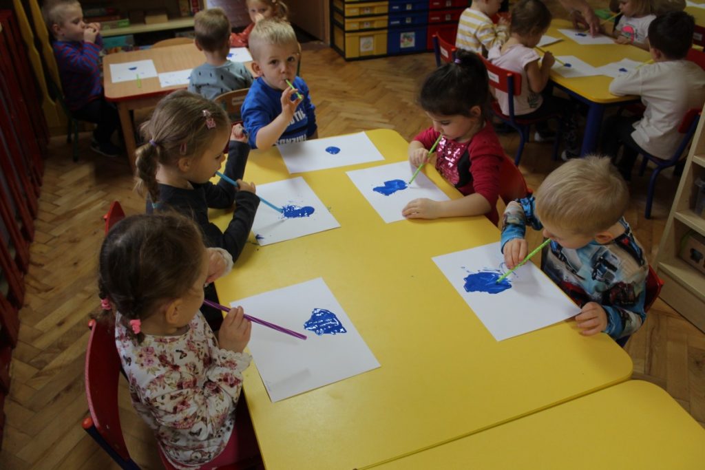 Dzieci przy stolikach dmuchają przez rurki na niebieskiego kleksa z farby