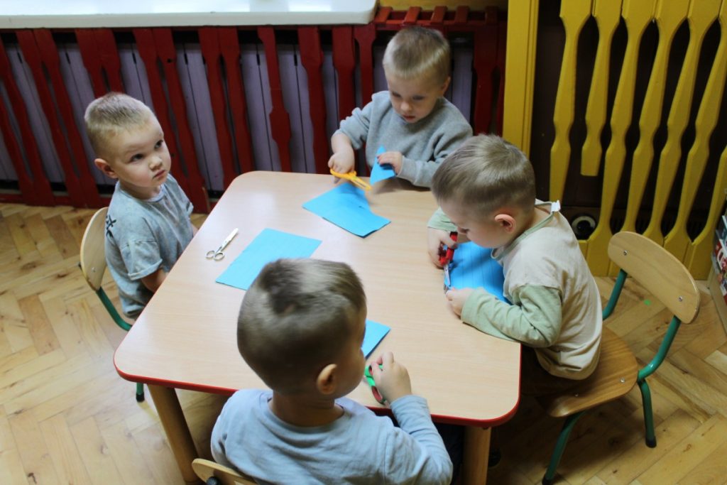 Dzieci przy stoliku wycinają nożyczkami paski niebieskiej kartki