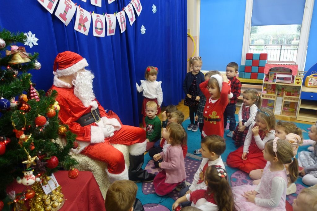 Dzieci stoją, siedzą na dywanie przed Mikołajem, który siedzi obok choinki