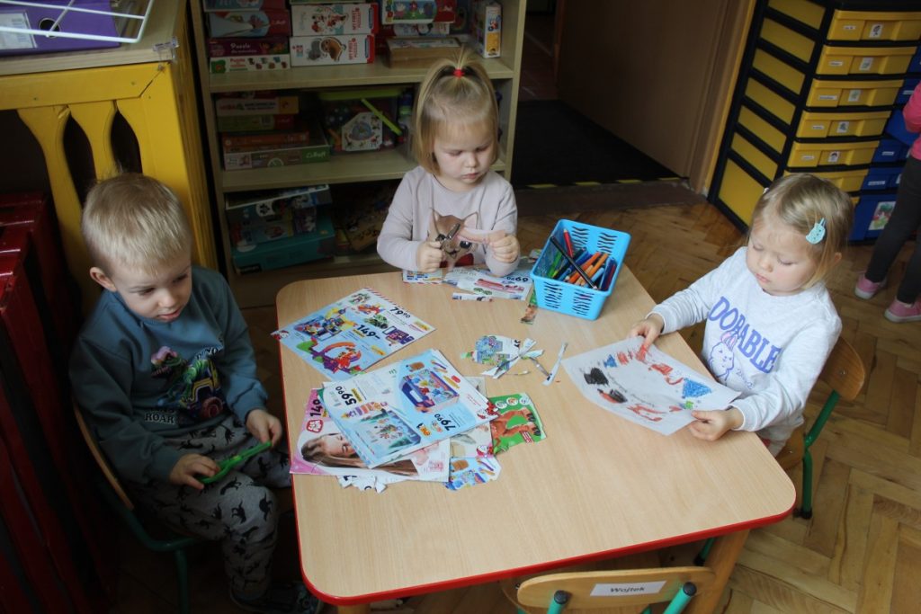 trójka dzieci przy stoliku wycina obrazki z gazet