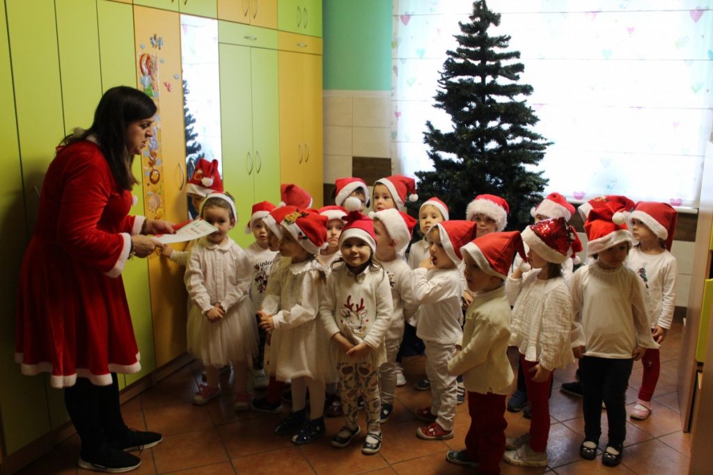 dzieci w białych strojach i czerwonych czapkach przy choince. Nauczycielka w stroju Mikołaja czyta list