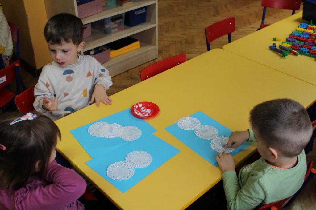 Trójka dzieci przy stoliku przykleja małe, białe kółeczka na niebieska kartkę