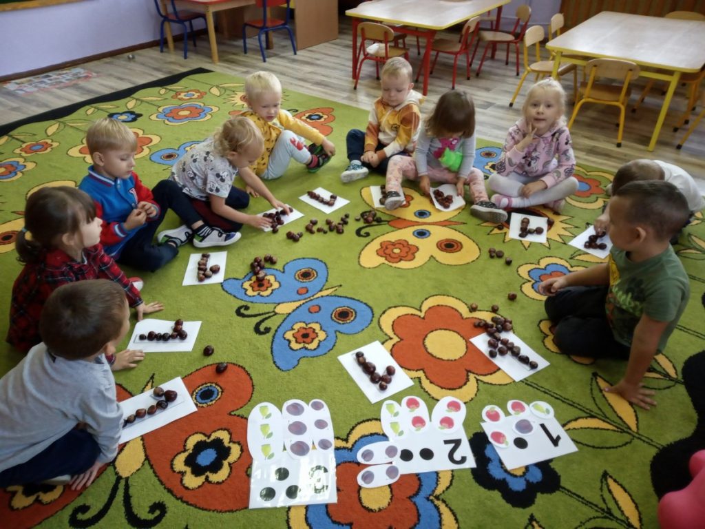 Na zdjęciu dzieci siedzą na dywanie, odwzorowują cyfrę 1 z kasztanów.