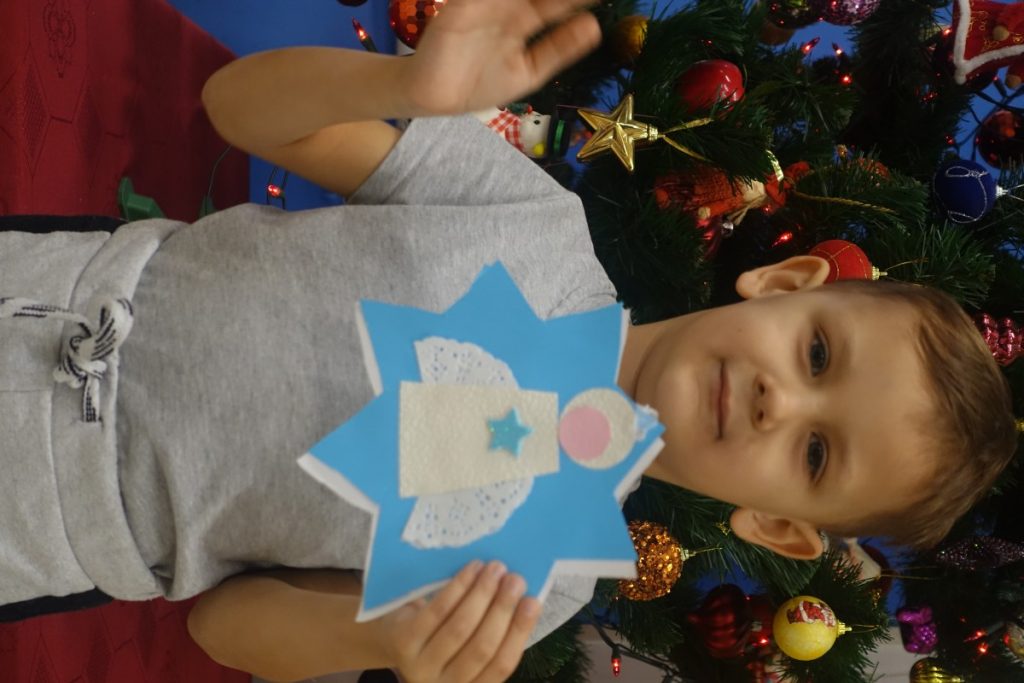 na zdjęciu dziecko stoi przed choinką, trzyma kartkę świąteczną 