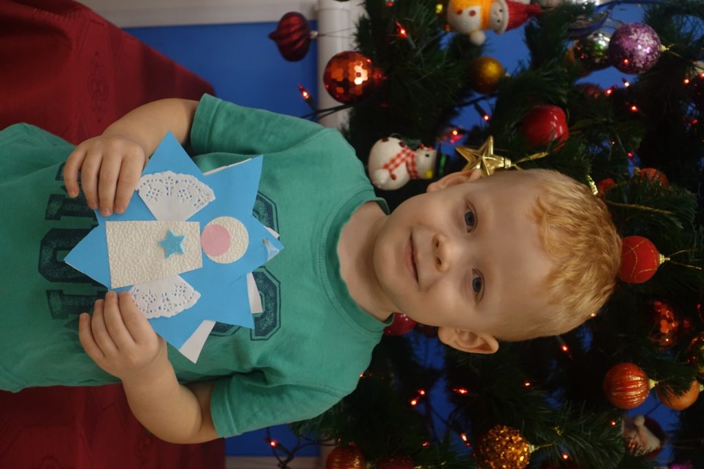 na zdjęciu dziecko stoi przed choinką, trzyma kartkę świąteczną 