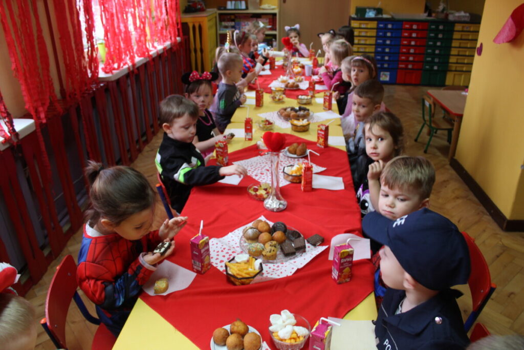 grupa dzieci siedzi przy stoliku przykrytym czerwonym obrusem