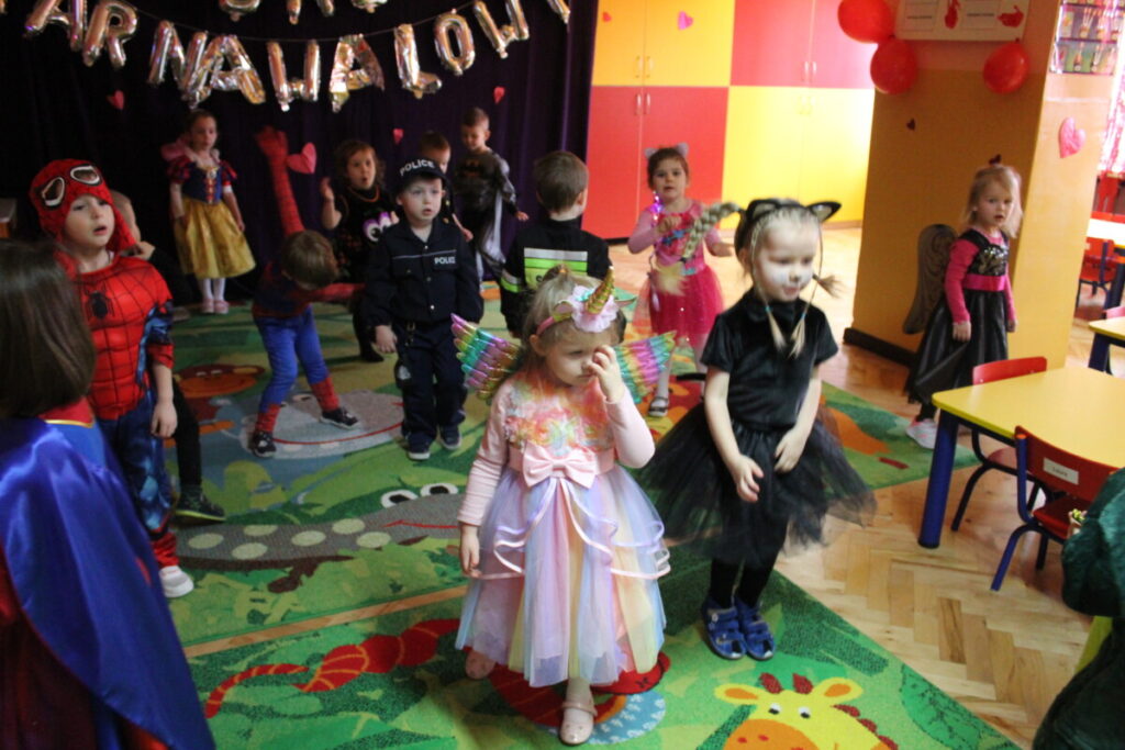 Dzieci tańczą na sali udekorowanej czerwonymi serduszkami w tle kotara z napisem bal karnawałowy