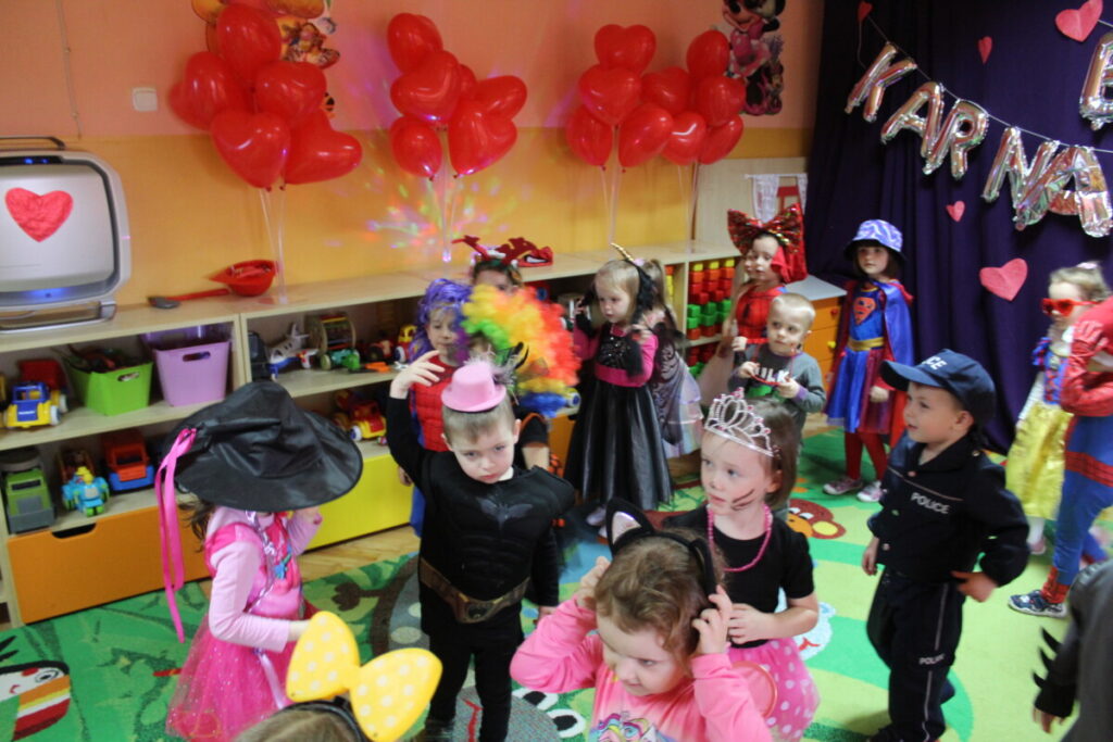 Dzieci tańczą na sali udekorowanej czerwonymi serduszkami w tle kotara z napisem bal karnawałowy