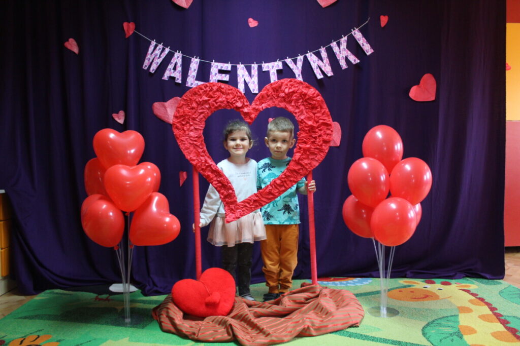 Para dzieci na ściance walentynkowej . W tle napis Walentynki po bokach czerwone balony w stojakach
