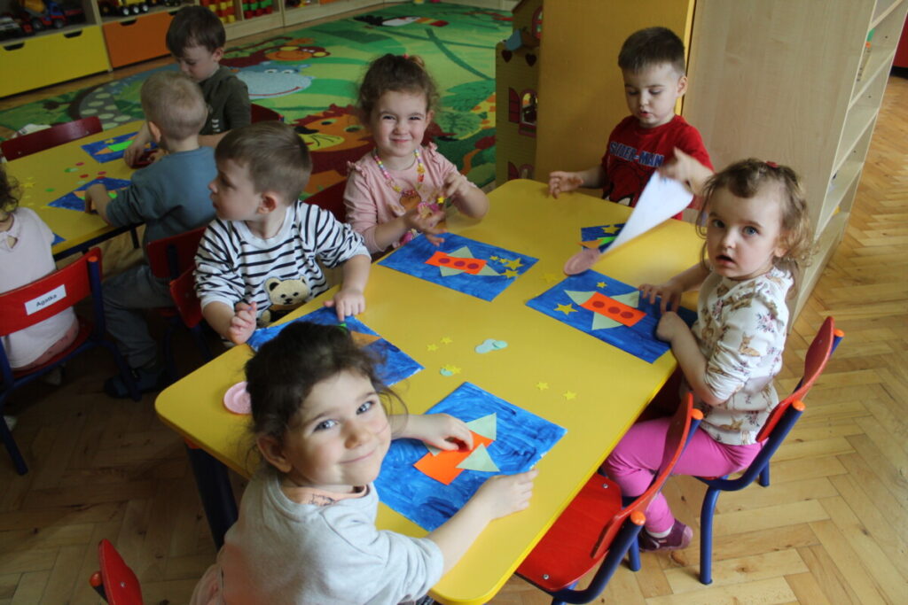 Dzieci przy stoliku przyklejają figury kolorowe na niebieskiej kartce