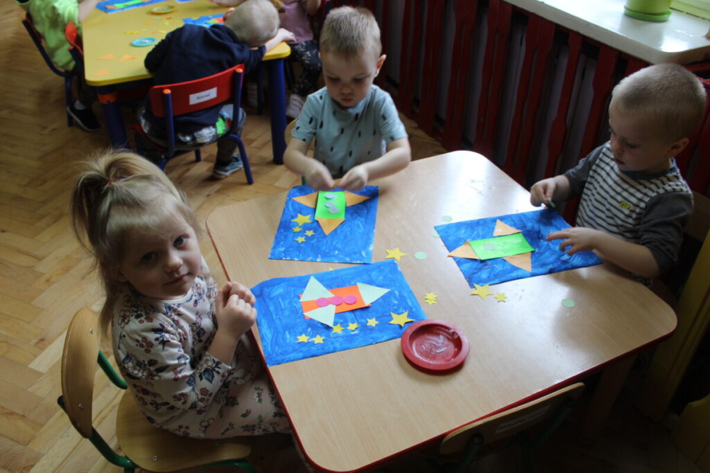 Dzieci przy stoliku przyklejają figury kolorowe na niebieskiej kartce