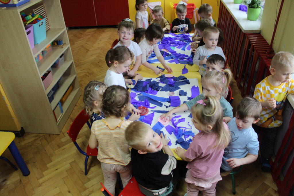 Dzieci przy stoliku wyklejają dużą kartkę niebieską bibułą. Praca w grupie