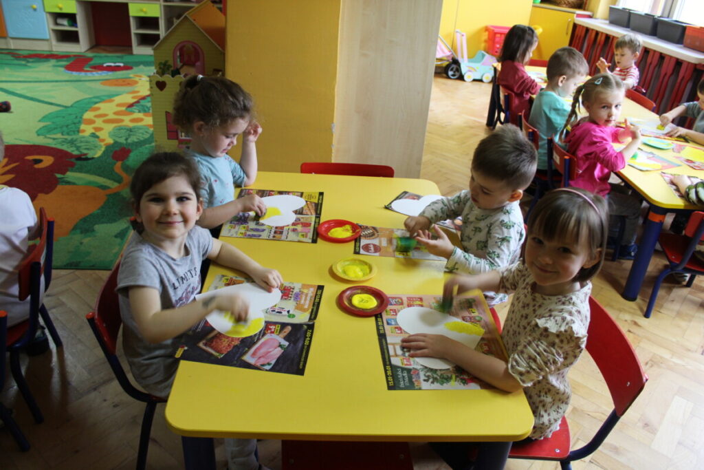 Dzieci przy stoliku malują żółtą farbą serduszka z papieru