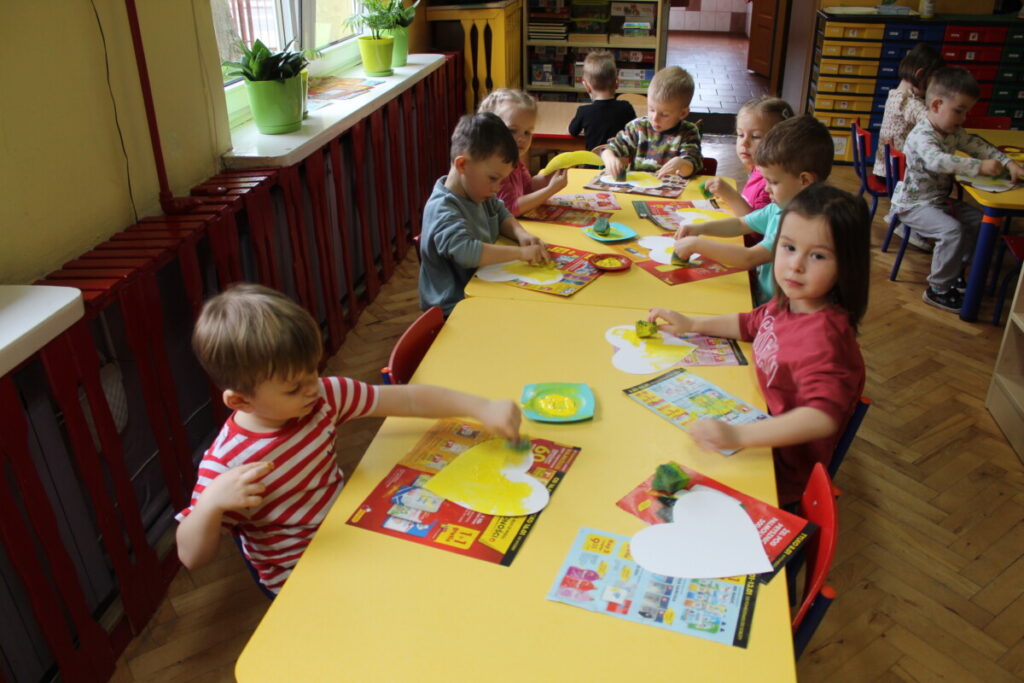 Dzieci przy stoliku malują żółtą farbą serduszka z papieru