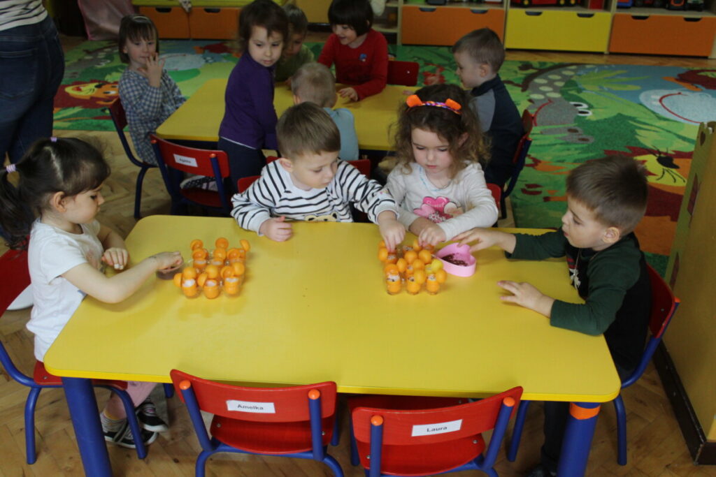 Dzieci przy stoliku sieją rzeżuchę w c czekoladowych po jajeczkach pojemnikach plastikowy