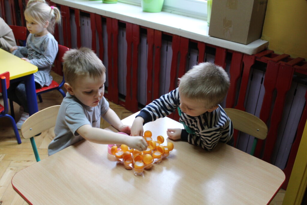 Dzieci przy stoliku sieją rzeżuchę w c czekoladowych po jajeczkach pojemnikach plastikowy
