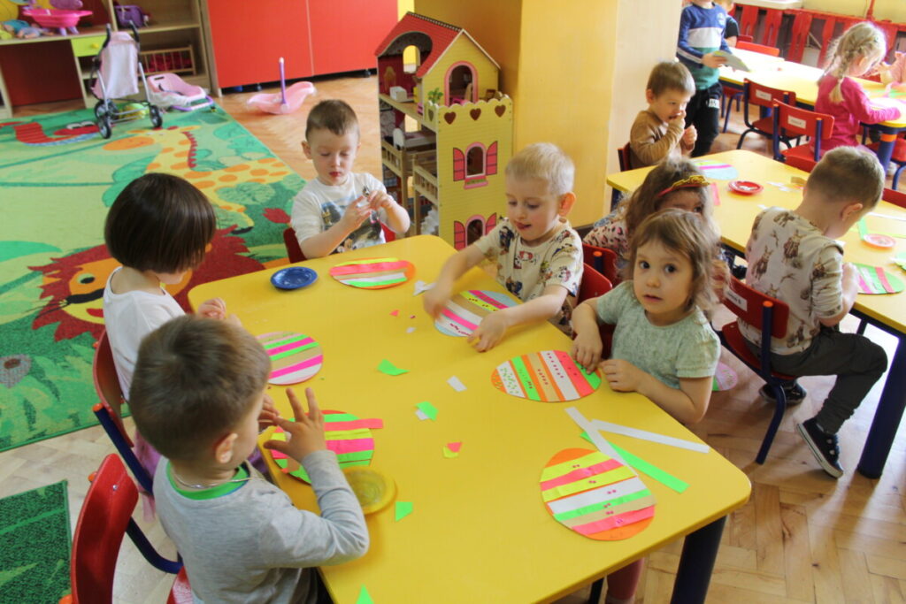 Dzieci przy stoliku wyklejają papierowe jajko kolorowymi paskami z papieru