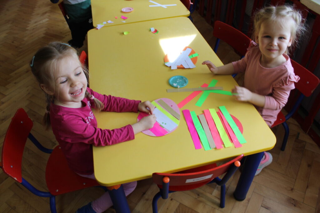 Dzieci przy stoliku wyklejają papierowe jajko kolorowymi paskami z papieru