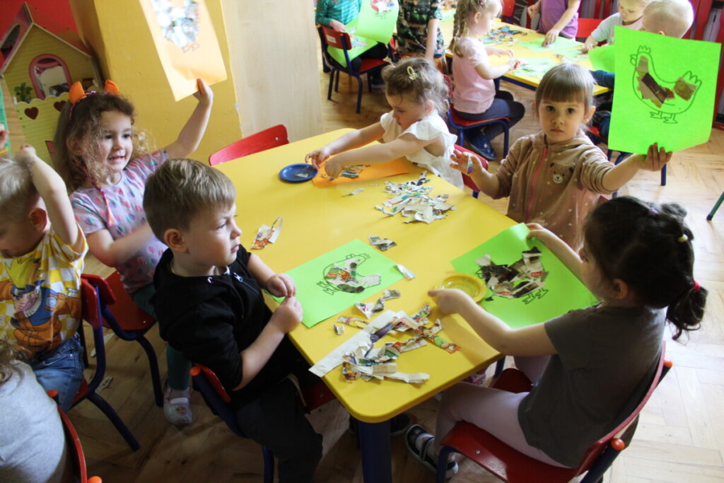 Grupa dzieci przy stoliku wykleja sylwetę kury kolorowymi kawałkami gazety