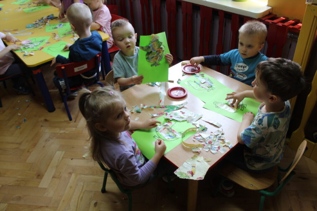 Grupa dzieci przy stoliku wykleja sylwetę kury kolorowymi kawałkami gazety