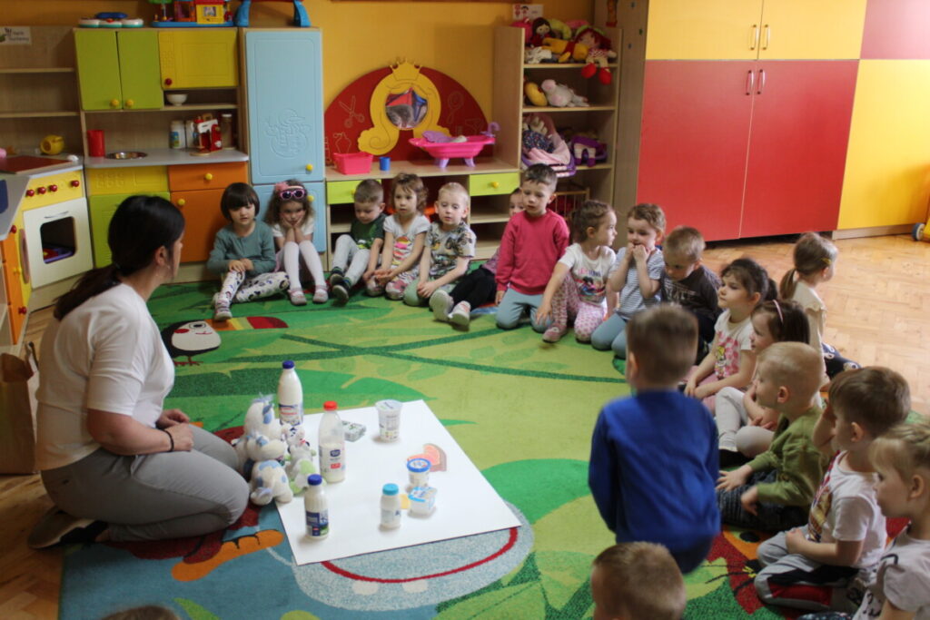 Dzieci na dywanie wraz z nauczycielka oglądają produkty które powstają z mleka