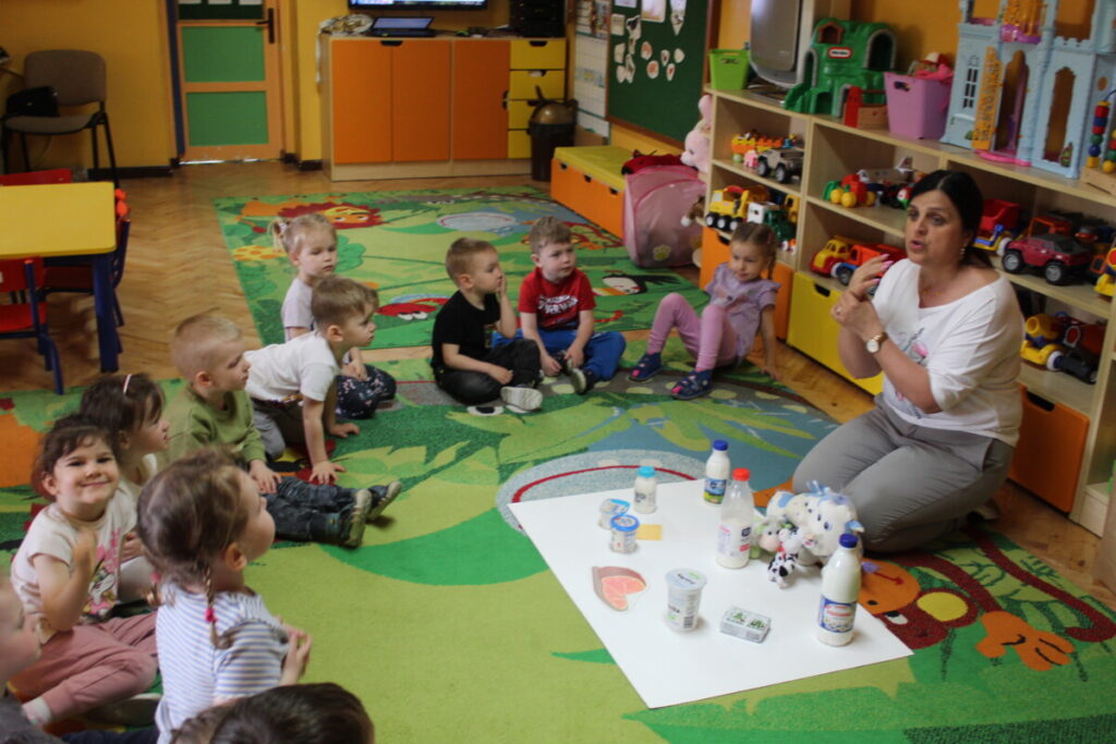 Dzieci na dywanie wraz z nauczycielka oglądają produkty które powstają z mleka