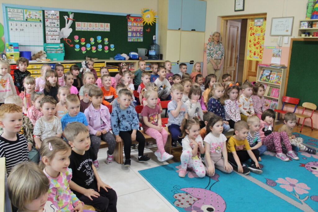 Grupa dzieci siedzi na krzesłach i na podłodze na sali przedszkolnej